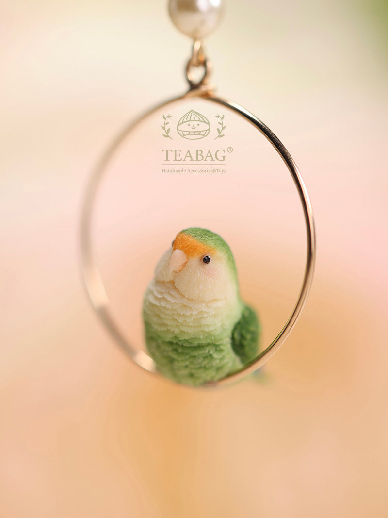 Portrait close-up of a green lovebird inside an earring hoop, showing its pale face, golden forehead, sculpted feathers and TEABAG branding.