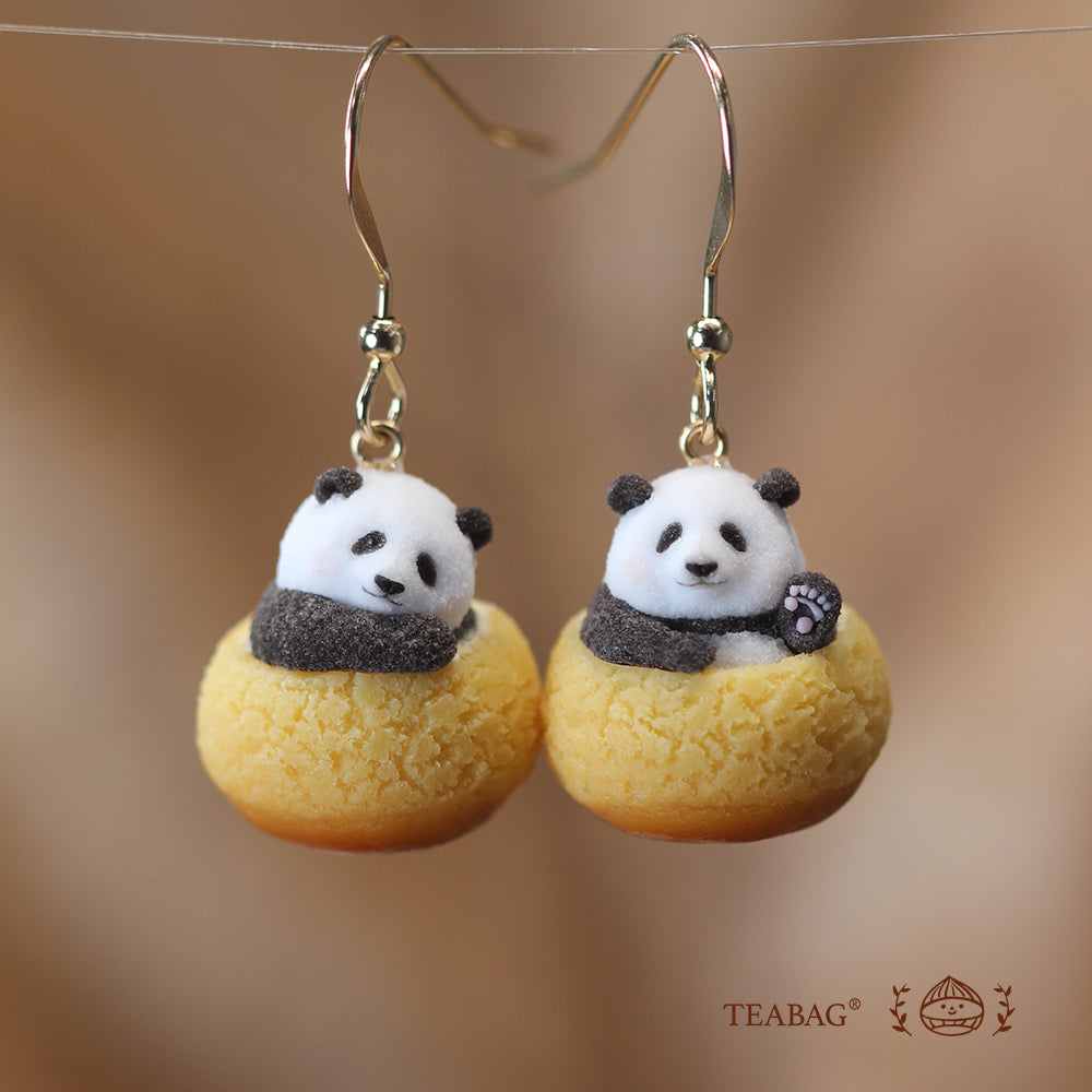 Pair of Panda Cream Puff hook earrings on a warm brown background, with one resting panda and one waving panda, showing the complete hooks and TEABAG branding.