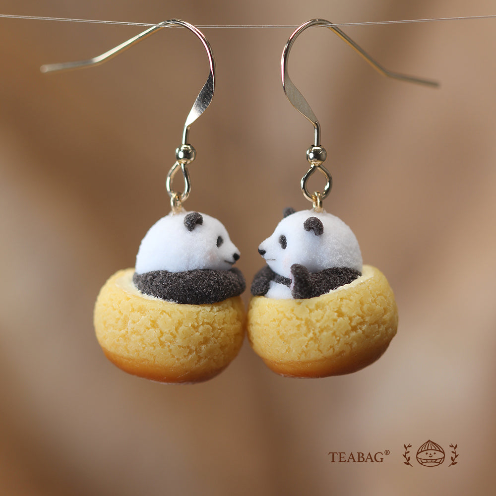 Side view of a pair of Panda Cream Puff hook earrings facing each other, showing both hooks and the cream puff texture against a warm brown background with TEABAG branding.