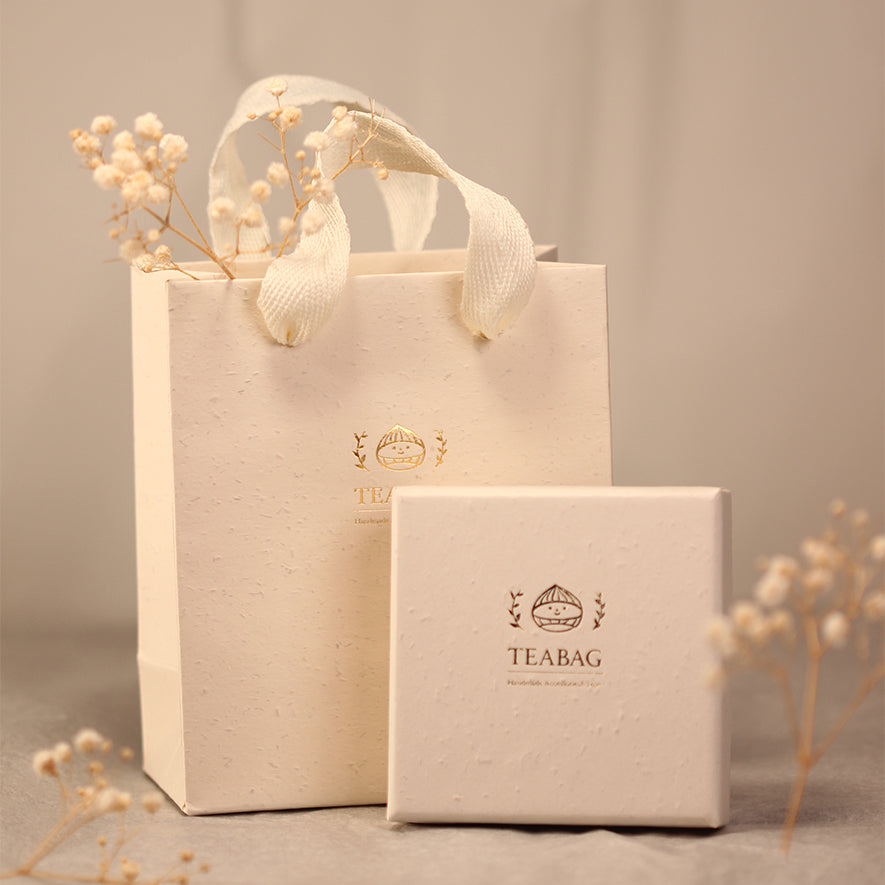 Cream TEABAG gift box and matching handled bag with gold branding, styled with dried flowers. Decorative flowers and styling props are not included.